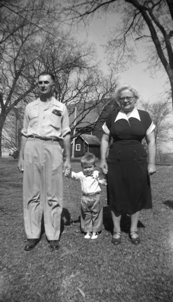 Glenn Oestrich standing between his grandparents, Edgar and Elna Krueger, holding their hands. In the background is the barn on the Krueger farm.