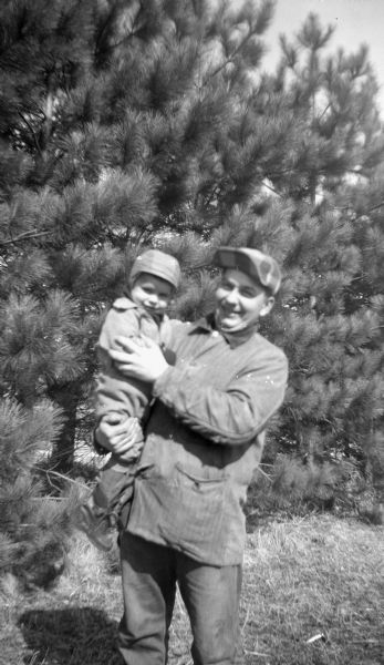 Robert Krueger holding his nephew Glenn Oestreich in his arms, in front of a row of pine trees.