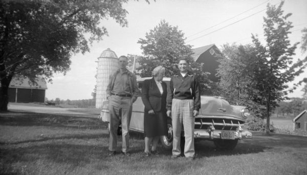Edgar, Elna, and Robert Krueger posing together in front of a Chevy automobile parked in their yard. The barn and a silo are in the background.