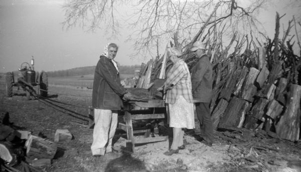 Elna Krueger stands with another woman and a man while cutting firewood using a belt-driven saw  by a tractor in a field in the background on the left. The women each hold a piece of wood on the saw table while the man watches. A pile of wood is stacked on the right.
