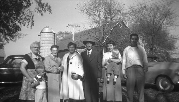 Group portrait in front of two cars parked on the Krueger farm. Elna Krueger stands on the far left with her grandson, Glenn Oestreich, standing in front of her. Robert Krueger stands on the far right next to his sister, Shirley Krueger Oestreich, who holds her son, Paul Oestreich. Two women and a man stand between Elna and Shirley. The barns and a silo are in the background.