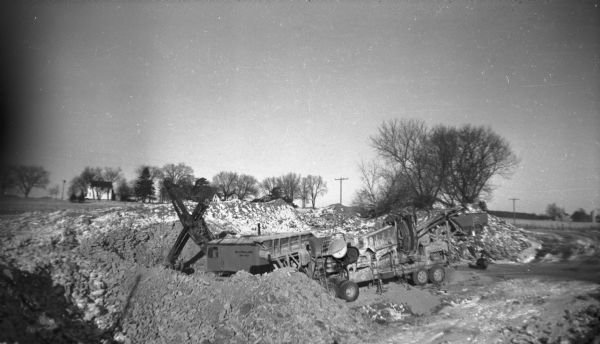 Slightly elevated view of machinery being used to dig up ground along a road near an open field. There is a farmhouse among trees in the background. Snow is on the ground.