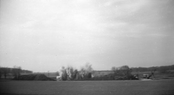 View across field of a dynamite explosion occurring between several mounds of dirt. A digger stands next to the dirt mound on the right.
