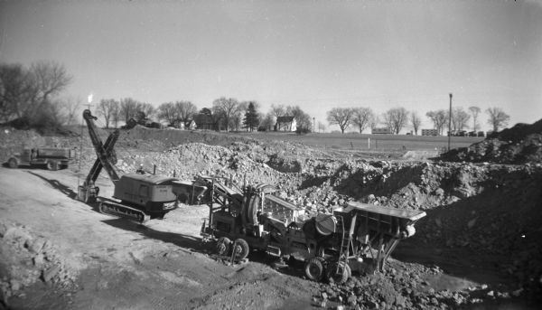 Slightly elevated view of a truck, crane and other digging equipment being used to dig up ground near a field. Several mounds of dirt are behind the machinery, and across the field in the background are farm buildings among trees.