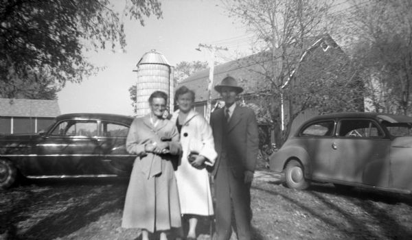 Group portrait of two women and a man standing together in front of two automobiles parked on the Krueger farm. In the background are barns and a silo.
