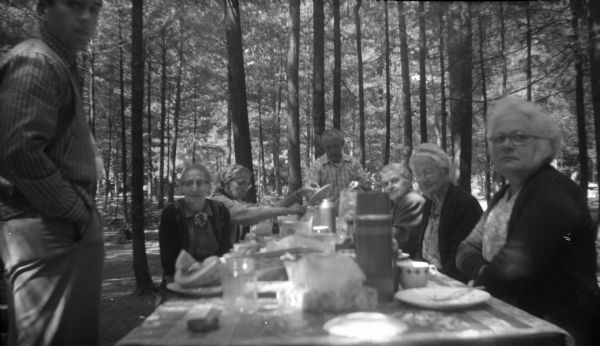 View from end of picnic table of five women and two men standing and sitting during a picnic in the middle of a woods. Plates, cups, thermoses, and various food items sit on the table. Elna Krueger sits on the end on the right side.