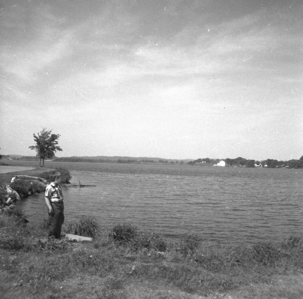 Glenn Oestreich standing along the shore of a lake. Another person sits on the curved shoreline in the background on the left. There are farm buildings on the far shoreline.