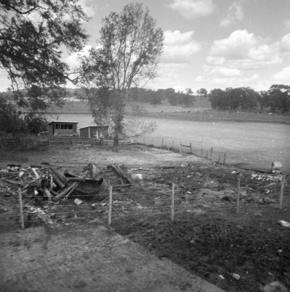 Slightly elevated view of debris littering a small fenced-in pen on the Krueger farm. The debris consists of a feeding trough, pieces of lumber, and other farm equipment. Cows are in a pasture on a low slope in the far background.