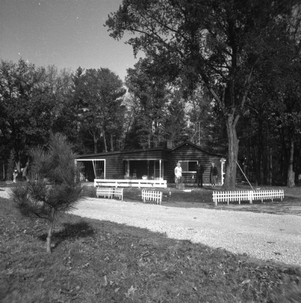 View across driveway towards Elna Krueger standing with a man and woman in the yard in front of a small cabin. Short white fences are along the driveway.