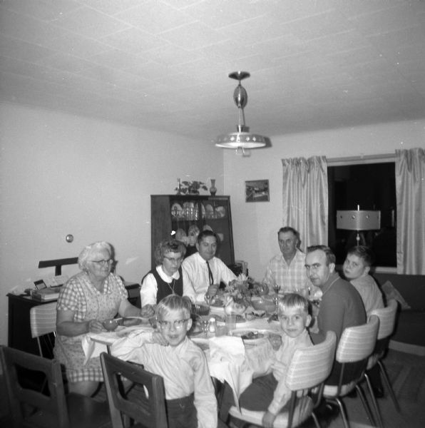 The Edgar Krueger family eating dinner together in the dinning room. A centerpiece of flowers inside a ceramic duck vase sit in the center of the table. Starting from the left and going clockwise: Elna Krueger, Shirley Krueger Oestreich, Robert Krueger, Edgar Kreuger, Glenn Oestreich, Delbert Oestreich, Dale Oestreich, and Paul Oestreich.