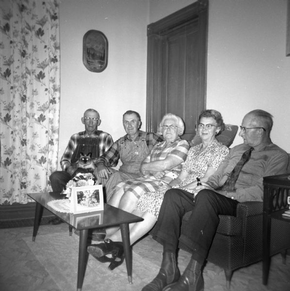 Edgar and Elna Krueger sitting with friends on a couch. A cat sits on the lap of a man in overalls in the center. Edgar sits in overalls next to him with Elna at his side.
