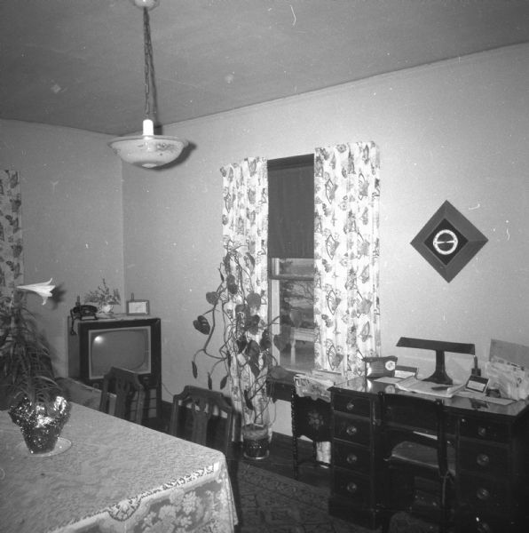 View of the dining room in the Krueger home. A television with a telephone on top stands in the corner behind the dining room table. A work desk and chair are next to the window on the right.
