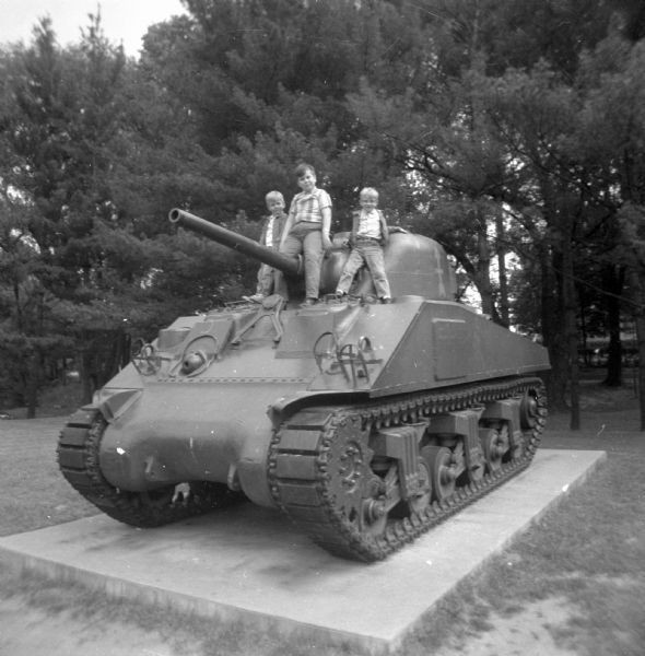 Dale, Glenn, and Paul Oestreich posing next to the turret of memorial Sherman Tank. A red arrow is painted on the tank's turret. In the background are trees.