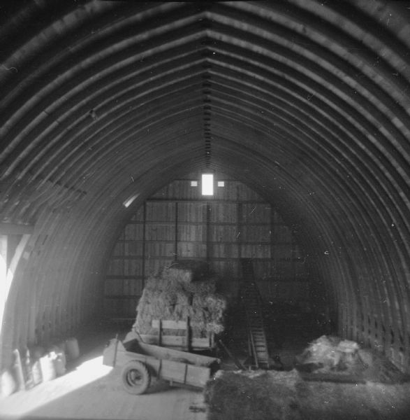 Elevated view from hayloft of the barn, with a wagon full of hay bales, an empty wagon, and a conveyer sitting in the center of the loft.