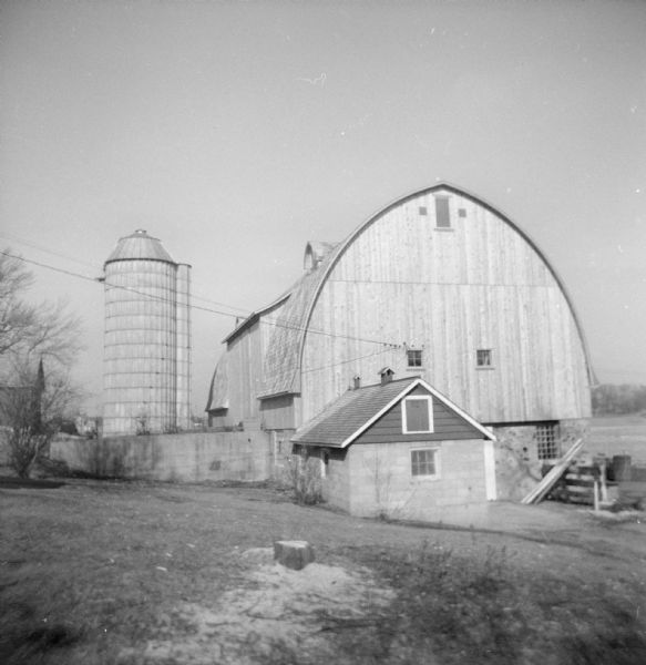 View down slope towards the new dairy barn, with a silo on the left. There is a tree stump with sawdust on the ground in the foreground.