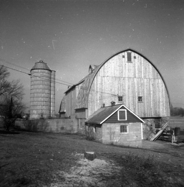 View down slope towards the new dairy barn, with a silo on the left. There is a tree stump with sawdust on the ground in the foreground.