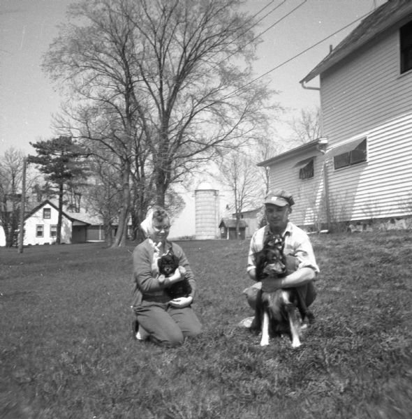 Portrait of couple kneeling on the lawn. The women on the left cradles a puppy in her lap, while the man holds a puppy in his right hand while his left arm is wrapped around the puppies mother. Farmhouses, a barn and a silo are in the background.