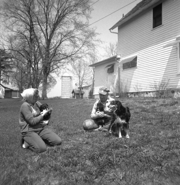 Portrait of a man and woman sitting on the lawn of the Krueger farm playing with puppies. Each of them is holding a puppy. The puppies mother stands next to the man on the right. A farmhouse, a silo, and several farm buildings are in the background.