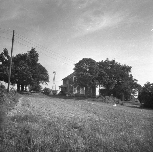 View across lawn towards a farmhouse obscured by a tree. A windmill is on the left next to the house, and farm buildings are in the background.