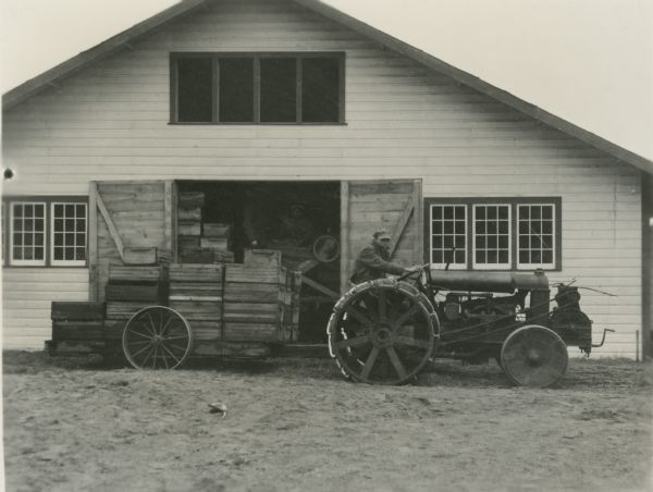A man driving a tractor is parked in front of a barn. The trailer on the back of the tractor is used for hauling crates of cranberries in from the fields during harvest. Two men stand inside the open doorway on a platform with a cleaning machine.
