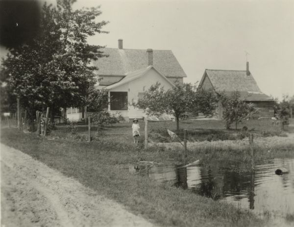 The farmhouse of the Wetherby Cranberry Company. At the time the farmhouse was owned by H. Kissinger. In the foreground is the corner of the water reservoir used to flood the cranberry marshes. A young girl is standing in the yard near a fence.
