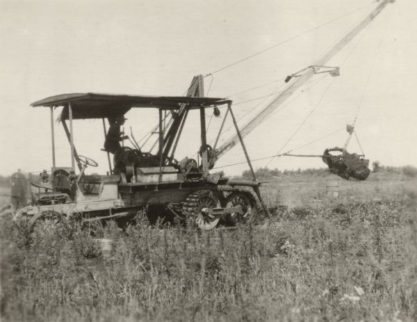 A man is sitting on the back of a Kissel truck using machinery to dredge a field. The field was being cleared in order to prepare it for use as a cranberry marsh.