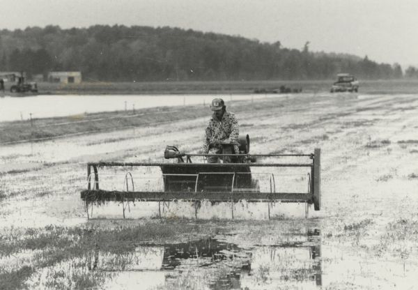 Man driving machine used to harvest cranberries on a flooded marsh. In the background is a truck and a tractor.