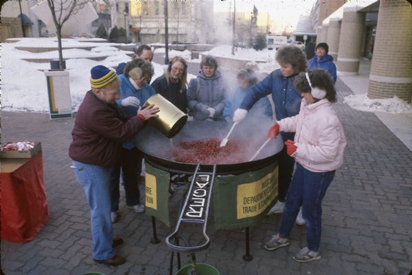 Outdoor gathering of men, women and children cooking cranberries in a large kettle, perhaps at a winter festival.