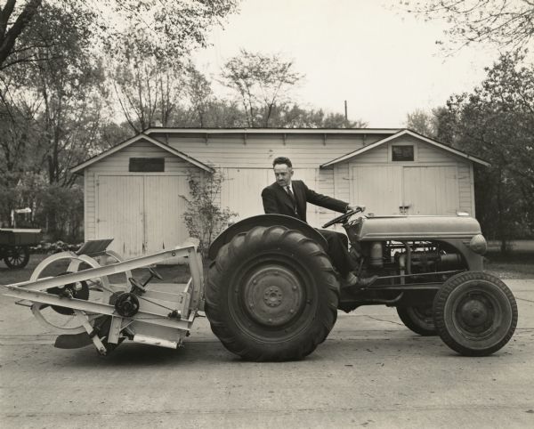 Farm implement attached to the back of a tractor driven by a man in a suit and tie. The machine is transported on the back of the tractor and then unloaded into a cranberry marsh. Once on the marsh the machine is used to rake the cranberries for harvest.