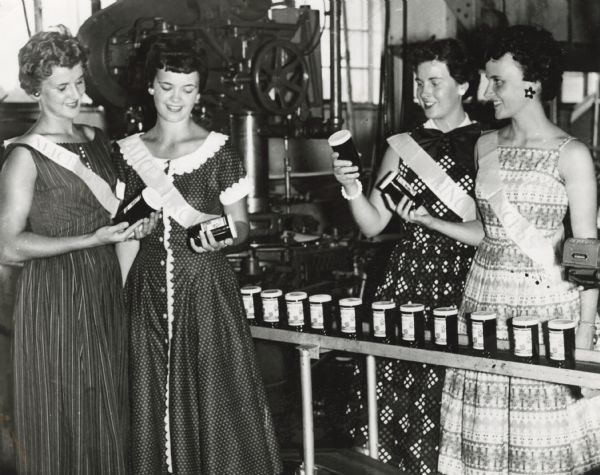 Alice in Dairyland and princesses inspect jars of cranberry sauce as they move along a production line.