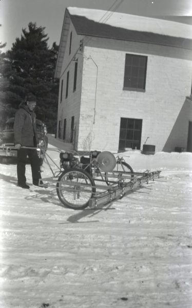Newell Jasperson of the Whittlesey Cranberry Company with machinery used in cranberry fields. There is snow on the ground, and behind him is a parked automobile and a large brick building.