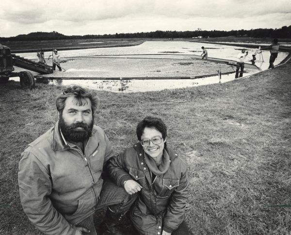 Tim Finch and Kay Finch of the Perry Creek Cranberry Company. In the background men work in the cranberry marsh to harvest the berries.