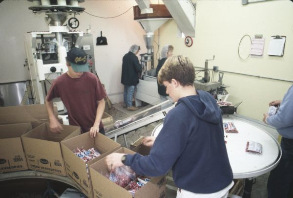 Two young men are packing boxes with bags of Ocean Spray cranberries. In the background women are bagging the cranberries.