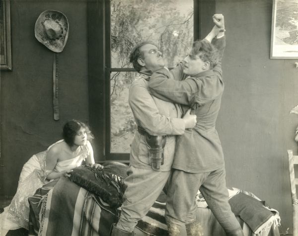 In dishabille, Paula Figueroa (played by Lenore Ulric) crouches on a bed in terror as Emiliano Pacheco (Howard Davies) and Bruce McLean (Forrest Stanley) fight. The scene is from the alternate happy ending of "The Heart of Paula," a silent drama of love and kidnapping set in Mexico.