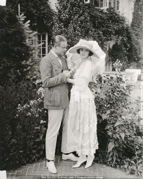 Huntly Gordon (in character as Harleth Crossey) and Irene Castle (as Marcia Crossey) pose outdoors in a full-length publicity photograph for "The Invisible Bond," her second silent film for Famous Players-Lasky. Irene Castle wears a beautiful long white dress, a large straw hat, and carries a parasol.