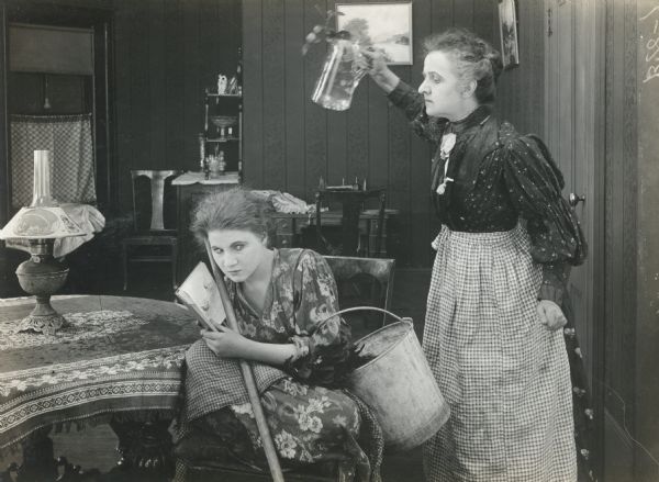 Florence Lawrence, seated at a table with a mop, bucket, and feather duster, has stopped work to read a book. Behind her an older woman, probably played by Laura Oakley, threatens her with a glass pitcher.