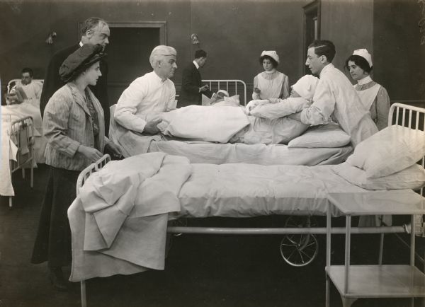 Florence Lawrence watches as a young man with bandaged hands and face is transferred by two orderlies from a gurney onto a hospital bed. Also visible in the scene still are other patients, nurses, and doctors in a hospital ward setting.