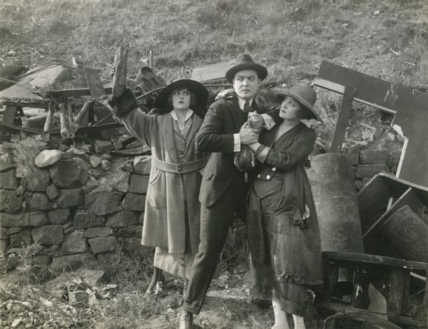 Pearl Date (played by Pearl White) threatens Captain Ralph Payne (Ralph Kellard) with a piece of firewood as he struggles with Bertha Bonn (Marie Wayne) for her purse in a publicity still from the 15-part silent serial "Pearl of the Army."