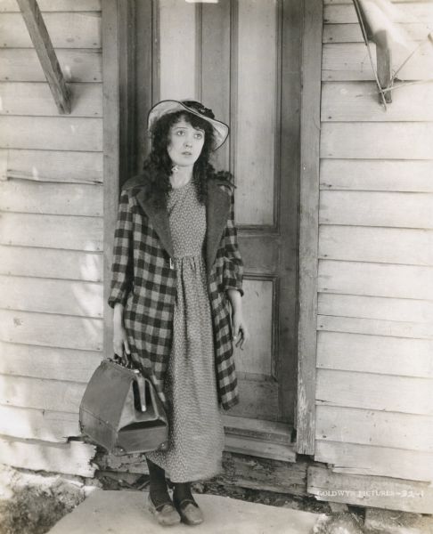 Minnie Penelope Peck (played by Mabel Normand) stands on the doorstep of an impoverished house with a traveling bag in her hand.