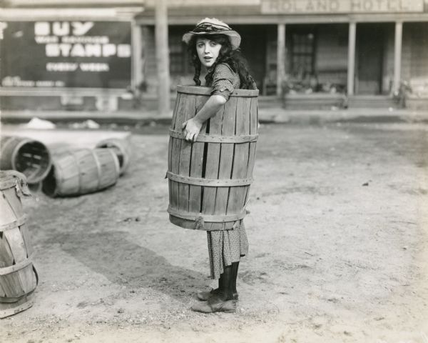 Minnie Penelope Peck (played by Mabel Normand) stands in the village street wearing a wooden barrel over the middle of her gingham dress. (Goldwyn 1918)