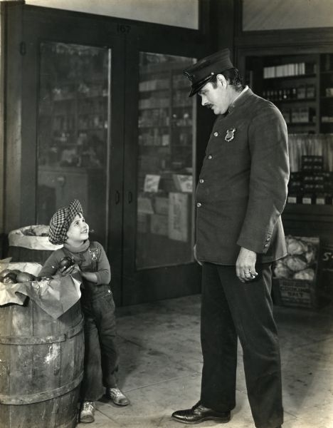 Henry Peck, the "bad boy" played by Jackie Coogan, is caught with his hand in the apple barrel by a police officer in this scene still from the silent film "Peck's Bad Boy" (First National 1921).