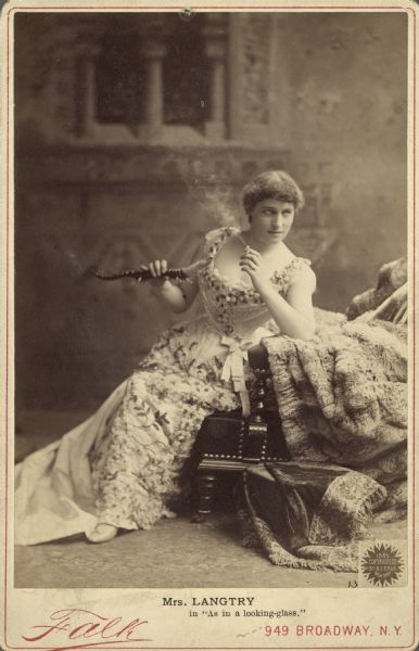 Lillie Langtry is in character as the adventuress Lena Despard from the play "As in a Looking-Glass." In this full-length portrait, she is  seated, facing right, holding a lizard-shaped cigarette lighter and smoking a cigarette in front of a painted backdrop.