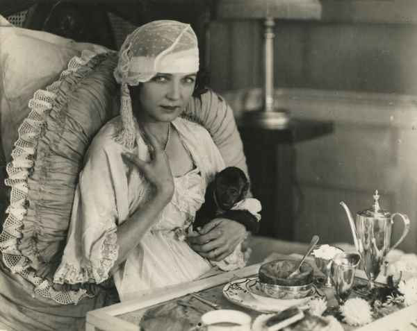 Irene Castle in pajamas and sleeping cap enjoying a cantaloupe as part of a posed breakfast in bed with her pet monkey Rastus. This photograph was used to promote the silent film "Patria."