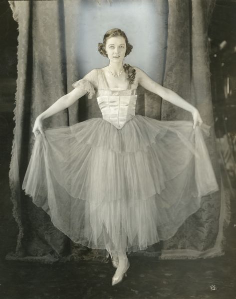 Irene Castle in a gauzy tulle and satin dress poses full-length in an air-brushed publicity photograph. The caption printed on the back of the print reads: "Mrs. Vernon Castle in the great preparedness motion-picture serial 'Patria.'"