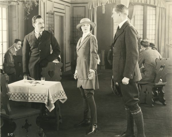 Cosmo Spotiswood (played by William P. Carleton on the right) confronts a man who has designs on his wife Justine (played by Irene Castle). Castle is dressed in horse-riding attire: hat, coat, jodhpurs, boots, and gloves. She holds a whip. This is a scene still from the 1920 Lasky production "The Amateur Wife."