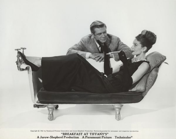 George Peppard (playing Paul Varjak) leans over the back in which lies Audrey Hepburn (as Holly Golightly) in a publicity still for "Breakfast at Tiffany's" (Paramount, 1961). Hepburn wears a sleeveless long black dress, elbow-length black gloves, a costume jewelry necklace, and smokes a cigarette in a long black holder. The sofa is an old cast-iron, claw-footed bathtub with one side removed.