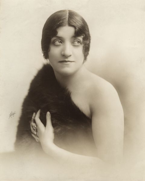 Half-length publicity photograph of Dorothy Jardon apparently wearing only a fur on one shoulder and many rings on her hand. Stamps on the back of the print suggest that the picture was distributed to promote a November 1915 production at B.F. Keith's Alhambra Theatre.