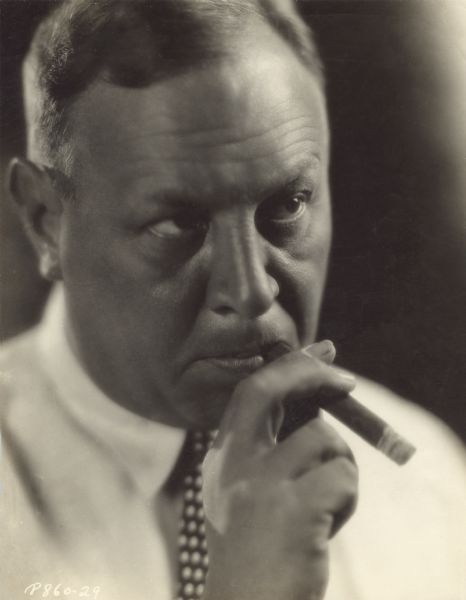 Close-up, low-key publicity portrait of Emil Jannings in shirt and tie holding a cigar to his mouth.