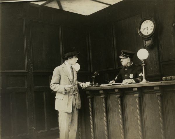 Jack Conway stands before the desk sergeant in a police station in a scene still from the one reel silent film "Added Fuel" (Reliance 1915).