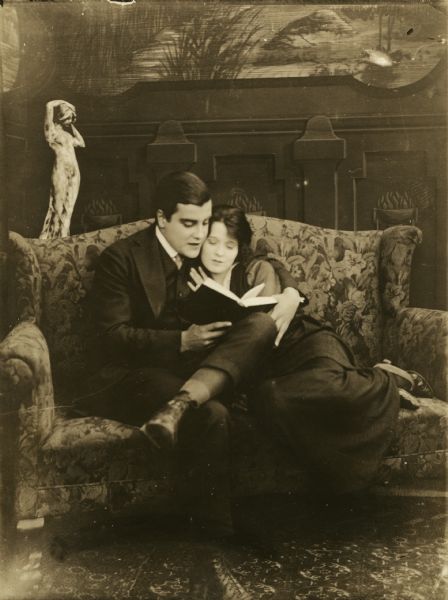 In a scene still from the one-reel silent film "Above Par," actors W.E. Lawrence and Billie West are curled up on a couch together reading a book. Lawrence plays Dick Carson, a newspaper reporter, who is the fiancé of a Wall Street broker's daughter.
